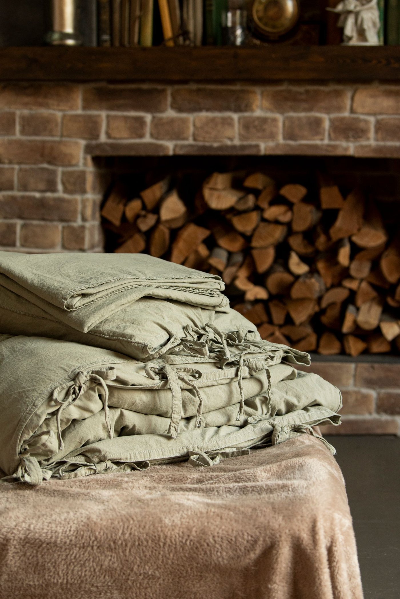 A cozy stack of folded sage-green bedding by a fireplace, ready for a seasonal linen refresh.